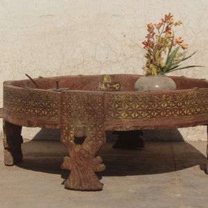 Pu&ograve; includere: Un tavolo rotondo in legno con intagli intricati e una finitura usurata. Il tavolo ha quattro gambe e un piano largo e piatto. Un piccolo vaso con fiori si trova sul tavolo.