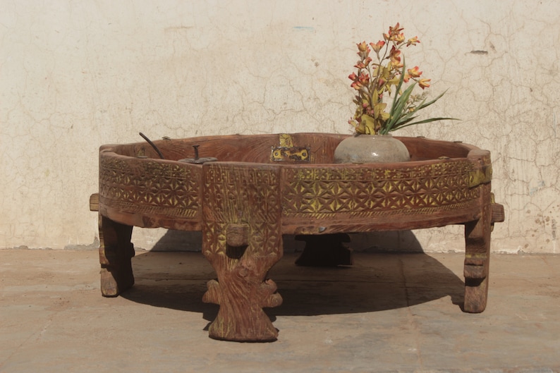 Pu&ograve; includere: Un tavolo rotondo in legno con intagli intricati e una finitura usurata. Il tavolo ha quattro gambe e un piano largo e piatto. Un piccolo vaso con fiori si trova sul tavolo.