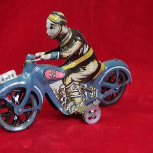 Könnte beinhalten: Vintage Spielzeugmotorrad zum Aufziehen mit Fahrer. Das Motorrad ist hellblau mit schwarzen Rädern und einem weißen vorderen Kotflügel mit der Nummer "F-22". Der Fahrer trägt Schwarz und Gelb mit weißem Gesicht und Helm.