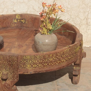 Pu&ograve; includere: Un tavolo rotondo in legno con un bordo intagliato e tre gambe. Il tavolo ha una finitura marrone chiaro ed &egrave; decorato con vernice gialla. Un piccolo vaso in ceramica grigia con fiori arancioni si trova sul tavolo.
