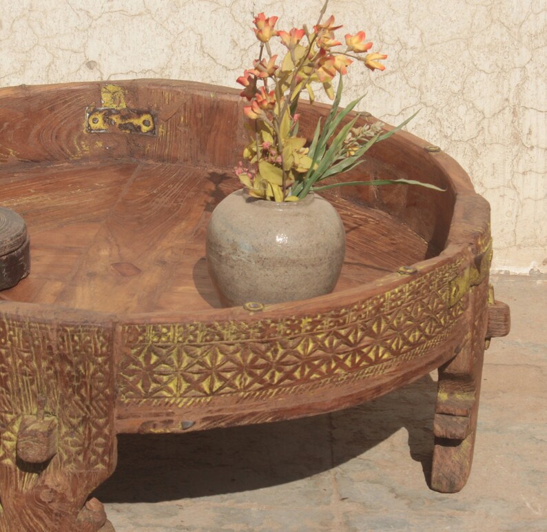 Pu&ograve; includere: Un tavolo rotondo in legno con un bordo intagliato e tre gambe. Il tavolo ha una finitura marrone chiaro ed &egrave; decorato con vernice gialla. Un piccolo vaso in ceramica grigia con fiori arancioni si trova sul tavolo.