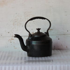 以下が含まれることがあります： 曲がった注ぎ口と大きなハンドルが付いた、ヴィンテージスタイルの黒い金属製ケトル。 ケトルは丸みを帯びたボディと小さなノブが付いた蓋が付いています。 白い木製の背景に対して、白い表面に置かれています。