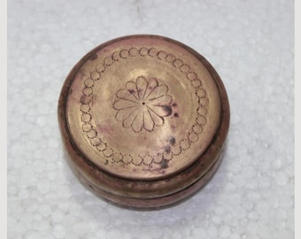 Pode incluir: Uma caixa redonda de latão antigo com um desenho floral na tampa. A tampa apresenta um motivo floral central rodeado por um círculo pontilhado. A caixa tem uma pátina quente e envelhecida, sugerindo sua natureza vintage. Cerca de 7 cm de diâmetro.