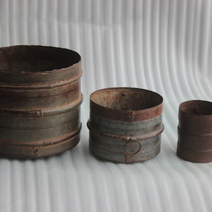 Könnte beinhalten: Drei rustikale Metallbehälter in verschiedenen Größen. Der größte Behälter ist dunkelgrau mit einer leicht rostigen Oberfläche und hat dekorative Bänder um die Ober- und Unterseite. Der mittlere Behälter ist in einem ähnlichen Grau mit einer rostigen Oberseite und einem kreisförmigen Design auf der Vorderseite. Der kleinste Behälter ist dunkelbraun mit einer rostigen Oberfläche.