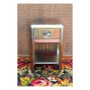 Puede incluir: Una pequeña mesa auxiliar de metal multicolor con un cajón y un estante inferior. La mesa está hecha de paneles de metal de cobre, plata y oro.