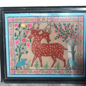 May include: A framed embroidery depicting two brown deer with white spots standing in a garden with green and pink flowers. The embroidery is on a beige background with a blue border and the text "KRM" in the top center.