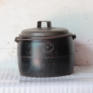 以下が含まれることがあります： 蓋付きの黒い鋳鉄製の鍋。 鍋には蓋にハンドルが1つ、側面にハンドルが2つ付いています。 鍋は白い表面に置かれています。 鍋の前面には円形のエンブレムが付いています。