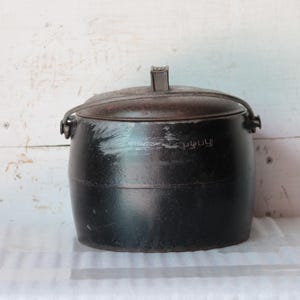 May include: A black, round cooking pot with a lid and handle. The pot has a dark, textured surface and a metal handle. The lid has a small handle on top. The pot is sitting on a white surface.