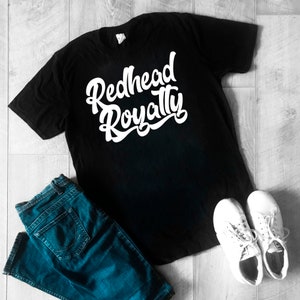 Op de afbeelding: Zwart T-shirt met witte tekst die "Redhead Royalty" luidt in een gestileerd lettertype.
