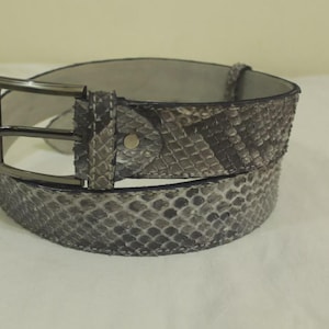 May include: A gray and black snakeskin belt with a rectangular gunmetal buckle. The belt is made of genuine snakeskin and has a textured pattern. The belt is a stylish accessory.