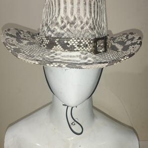 Può includere: Un cappello da cowboy in pelle di serpente bianco e grigio con una fascia in pelle marrone e una fibbia argentata. Il cappello è su una testa di manichino bianca.