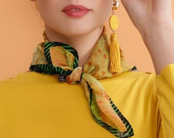 May include: A woman wearing yellow round sunglasses with dark lenses, a yellow top, and a patterned scarf. The scarf is tied around the neck. The woman is also wearing yellow earrings with a tassel.