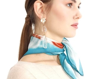 May include: A woman wearing a light blue and white patterned silk scarf tied around her neck. She is also wearing long, clear, teardrop-shaped earrings and a beige sweater. The scarf has orange accents.