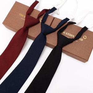 May include: Three clip-on ties in burgundy, navy blue, and black. The ties are made of a smooth fabric and have a simple design. The ties are displayed on a brown box with gold lettering that reads "VOCATION BRAND".