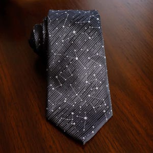 May include: A dark grey necktie featuring a repeating white pattern of celestial constellations and stars. The fabric has a subtle diagonal ribbed texture throughout, with the constellation lines and star points printed in a crisp white ink.