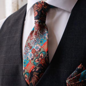 May include: A patterned necktie with a matching pocket square. The tie features a mix of floral and paisley designs in shades of red, blue, and brown against a black background. The tie is knotted and worn with a white dress shirt and a dark gray vest.