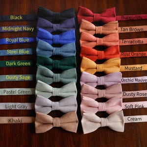 Peut inclure: Une collection de nœuds papillon de différentes couleurs, dont le noir, le marine, le bleu et le rose. Chaque nœud papillon est présenté avec un ruban assorti, avec des noms de couleurs comme "Bourgogne" et "Moutarde" visibles.