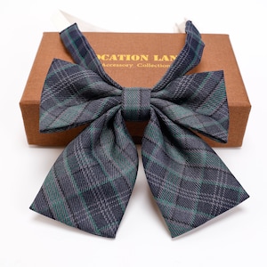 Può includere: Un papillon a quadri nei toni del blu navy, verde e grigio, con una forma a farfalla classica. Il papillon è presentato su una scatola marrone con la scritta "LOCATION LANE Accessory Collection". Il papillon è realizzato in tessuto.
