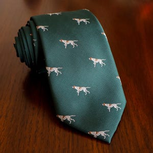 May include: A dark green necktie featuring a repeating pattern of small white and brown hunting dogs in a running pose. The fabric has a visible diagonal ribbed texture throughout the entire length of the tie, which is rolled at the top end.