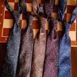 May include: Five neckties arranged side by side, each featuring a diagonal weave texture and a white constellation pattern. The colours from left to right are black, dark brown, medium brown, deep burgundy, and navy blue, all tied in knots.