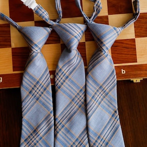 May include: Three pre-tied neckties featuring a light blue, white, and tan diagonal plaid pattern. Each tie includes a white plastic adjustable clip mechanism at the top, designed for easy attachment, with a structured knot and a tapered blade shape.