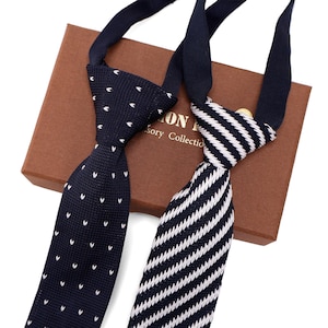 Könnte beinhalten: Zwei Krawatten mit verschiedenen Mustern. Eine ist marineblau mit kleinen weißen Punkten, die andere hat weiß-marineblaue Streifen. Die Krawatten liegen auf einer braunen Schachtel mit dem Text "ON Accessory Collection".