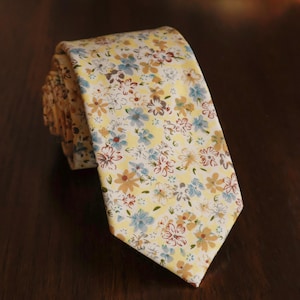 May include: A necktie featuring a light yellow background with a repeating floral pattern in shades of light blue, tan, and dark brown. The tie is rolled at the top and tapers to a pointed end, displaying a dense arrangement of small flowers.