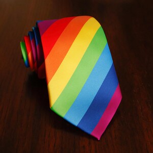 May include: A necktie featuring a vibrant diagonal rainbow stripe pattern. The colours transition from red, orange, and yellow to green, blue, and purple. The tie is rolled at the top, showing the layered fabric folds of the smooth, satin-like material.