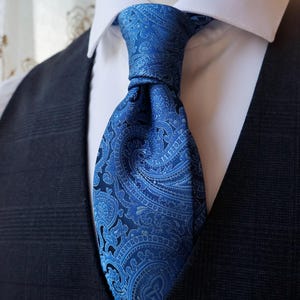 Peut inclure: Une cravate bleue ornée d'un motif cachemire ton sur ton dans des nuances de bleu variées. La cravate est nouée et portée sur une chemise blanche à col rigide, sous un gilet à carreaux gris anthracite foncé avec une finition texturée.