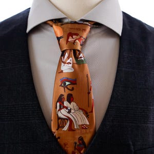 Könnte beinhalten: Eine braune Seidenkrawatte mit altägyptischen Hieroglyphen und Figuren. Die Krawatte ist geknotet und wird mit einem weißen Hemd und einer dunkelblauen Anzugjacke getragen. Das Design der Krawatte zeigt sitzende Figuren und das Horusauge.