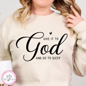 May include: A beige sweatshirt with the text "GIVE IT TO GOD AND GO TO SLEEP" in black cursive lettering. A small heart is above the word "GOD".
