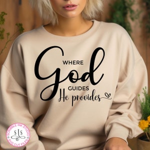 Puede incluir: Una sudadera beige con el texto "Where God Guides He Provides" en letras cursivas negras con un pequeño símbolo de corazón.