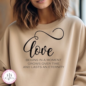 Könnte beinhalten: Ein beiger Pullover mit dem Text "love begins in a moment grows over time and lasts an eternity" in schwarzer Schrift. Das Wort "love" ist in einer kursiven Schrift.