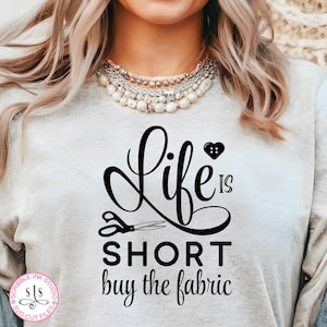 Puede incluir: Una camiseta blanca con texto negro que dice "Life is short buy the fabric". El texto está en una fuente cursiva e incluye un pequeño corazón con un botón en el centro. Hay tijeras debajo del texto.