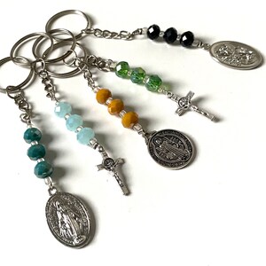 Pode incluir: Cinco chaveiros prateados com contas e pingentes de diferentes cores. Cada chaveiro tem uma cruz prateada, um pingente oval prateado com uma imagem religiosa e um anel de chave prateado. As contas são pretas, verdes, amarelas e azuis.