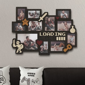 Puede incluir: Un marco de fotos de madera negro con un collage de fotos y gráficos de videojuegos pixelados. El marco tiene forma de consola de videojuegos e incluye una barra de carga, una estrella, una espada, una llave y una poción. El marco está colgado en una pared blanca sobre un sofá negro con dos almohadas blancas que presentan gráficos de controladores de videojuegos.
