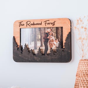 May include: Wooden photo frame with a natural wood finish and a dark brown forest silhouette design. The text "The Redwood Forest" is engraved at the top. The frame holds a photograph of a family in a forest setting.