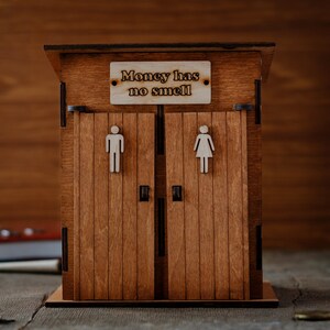 Puede incluir: Una caja de madera para guardar dinero con forma de letrina con dos puertas. El letrero sobre las puertas dice "El dinero no huele."