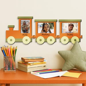 Op de afbeelding: Een oranje fotolijst in de vorm van een trein met drie wagons, elk met een foto. Het frame is aan een muur bevestigd boven een tafel met een stervormig kussen, boeken en kleurpotloden. De wagons zijn gelabeld met "Andy", "Mommy" en "Daddy".