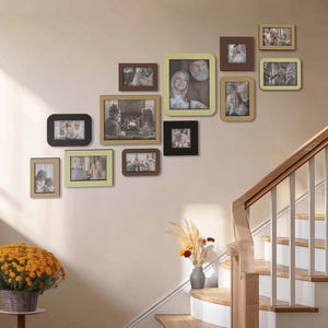 May include: A collection of framed photographs in various sizes and shapes, mounted on a light-colored wall. The frames are in shades of brown, black, and cream, with some featuring a yellow border. A staircase with wooden steps and a white railing is visible.