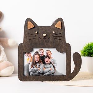 Puede incluir: Un marco de fotos de madera marrón oscuro con forma de gato con una foto de cinco niños. El marco tiene cola y orejas con detalles en marrón claro. El marco está sobre una superficie blanca con un peluche y una pequeña planta.
