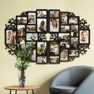 Puede incluir: Un marco de fotos de madera negro en forma de árbol con múltiples aberturas para fotos. El marco está decorado con recortes en forma de hojas. El marco está lleno de fotos de personas, algunas en grupo y otras solas. El marco está colgado en una pared de color beige claro.