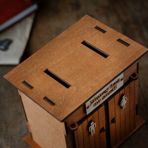 Puede incluir: Caja de madera para guardar dinero con forma de letrina con una ranura en el techo. La caja tiene un letrero que dice "El dinero no huele".