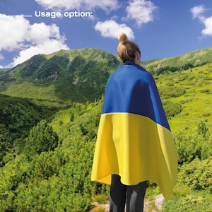 May include: A person standing in a mountainous landscape, wearing a blue and yellow flag draped over their shoulders. The flag is the Ukrainian flag.