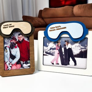 May include: Two wooden photo frames with ski goggles design. One frame is brown with the text "LOVE AT FIRST SNOW" and the other is white with the text "COLD DAYS, WARM MEMORIES". Each frame holds a photograph.