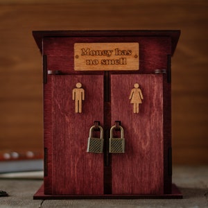 May include: A wooden box with two doors, each with a lock and a small cutout of a person. The box is painted a deep red colour and has the words "Money has no smell" written on the front.