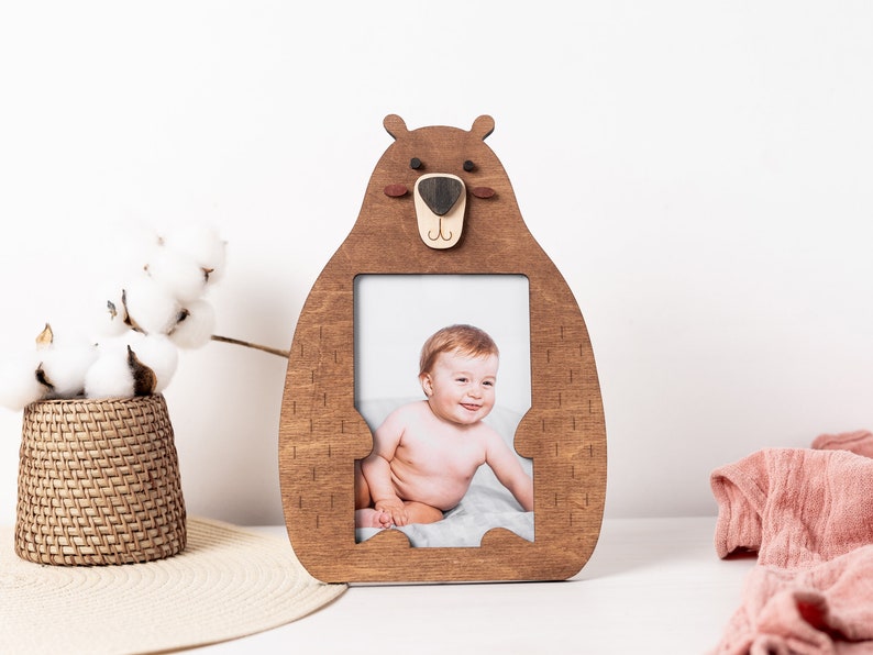 Puede incluir: Un marco de fotos de madera con forma de oso marr&oacute;n con un recorte para una foto. El oso tiene una nariz marr&oacute;n, ojos negros y una peque&ntilde;a sonrisa. Una foto de un beb&eacute; est&aacute; en el marco.
