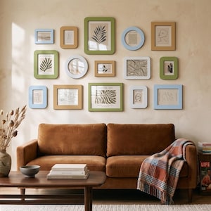 Op de afbeelding: Een galerijwand met vijftien verschillende fotolijsten in groen-, blauw- en bruintinten, met diverse lijntekeningen. De rechthoekige en ronde lijsten zijn bevestigd aan een muur boven een bruine fluwelen bank.