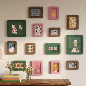 Puede incluir: Una colección de obras de arte enmarcadas en varios tamaños y colores, dispuestas en una pared blanca. Los marcos son marrones, verdes y rosas, con imágenes de gatos, pájaros y otros diseños caprichosos. La palabra "COOL" se muestra en un marco.