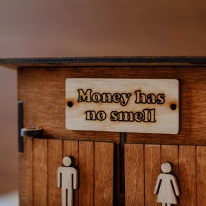 Puede incluir: Una caja de madera con dos puertas, cada una con la silueta de una persona. La caja tiene un letrero que dice "El dinero no huele."